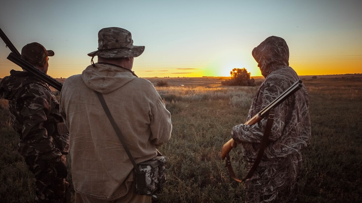 Three hunters standing shoulder to shoulder at sunset, seen from behind, looking out over an open field.