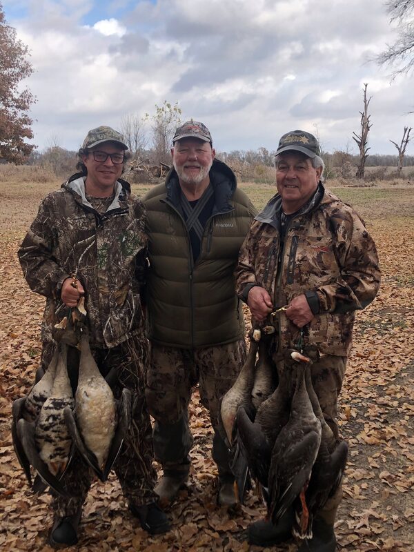 Three hunters standing shoulder to shoulder in fallen leaves, each holding the geese they took that morning.